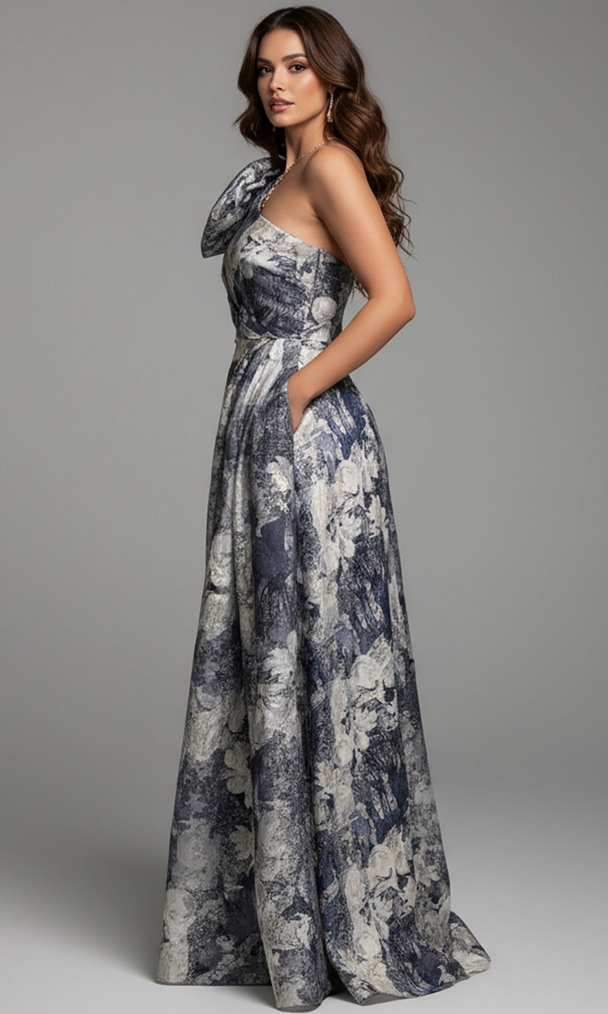 A woman wears the Jovani 25663 long formal dress featuring a sleeveless, asymmetrical neckline, side pockets, and an a-line skirt in a blue and white floral print against a plain gray background.